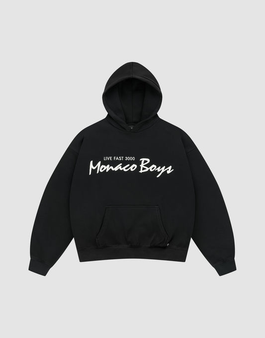 The Monaco Boys Hooded by LIVE FAST DIE YOUNG is a black oversized hoodie with a front pocket, featuring "LIVE FAST 3000 Monaco Boys" printed in white on the chest and finished with a subtle flag-label detail. | plain