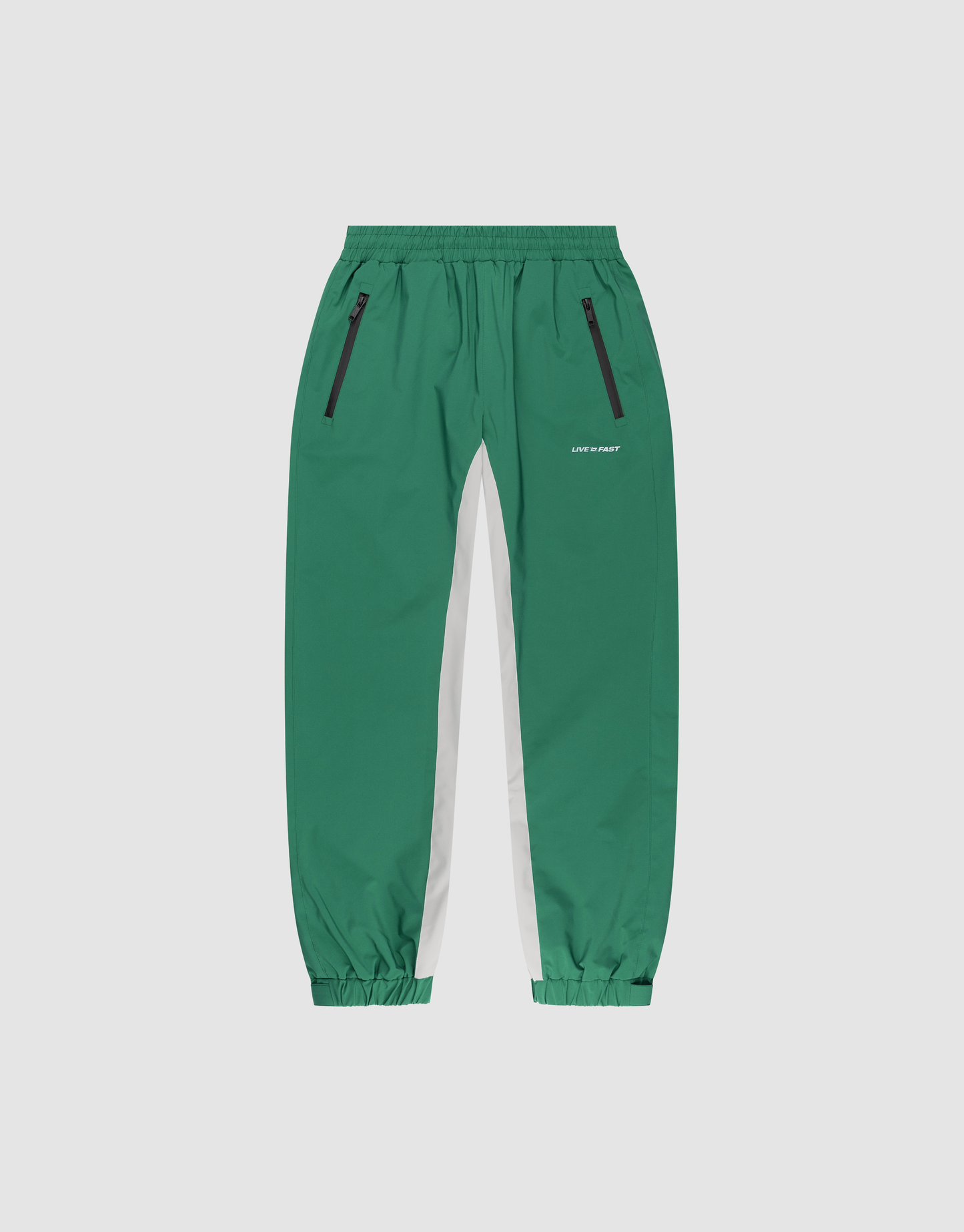 Basic Shell Pants by LIVE FAST DIE YOUNG: green track pants with elastic waistband and cuffs, black zippered side pockets, a white inner leg stripe, and small reflective "LVR FIRST" text on the left thigh, shown on a plain background.