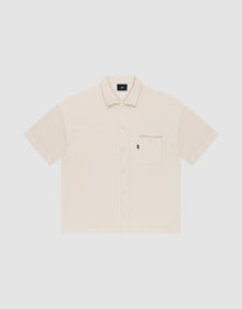 The Basic Knit Shirt by LIVE FAST DIE YOUNG is a short-sleeve, light beige button-up with a Hemdkragen, front chest pocket, and small black LFDY branding tag on the pocket, shown on a plain white background. | plain