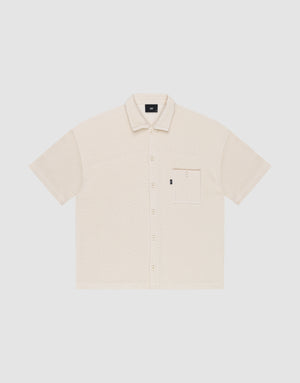 The Basic Knit Shirt by LIVE FAST DIE YOUNG is a short-sleeve, light beige button-up with a Hemdkragen, front chest pocket, and small black LFDY branding tag on the pocket, shown on a plain white background. | plain