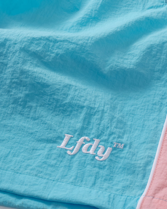 Close-up of light blue Beach Shorts by LIVE FAST DIE YOUNG, featuring LfdY™ embroidered in white and a pink and white edge stripe—regular fit for a stylish beachwear look.