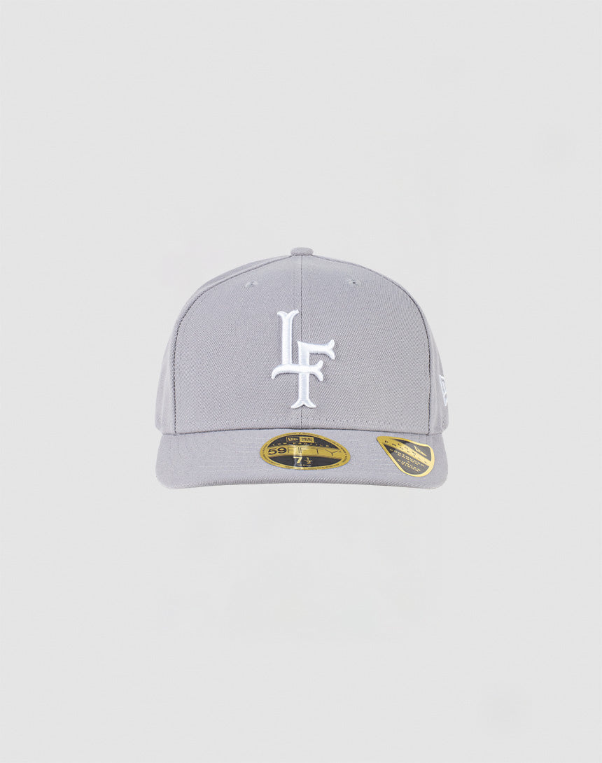 The LFDY x NEW ERA 59FIFTY Low Profile cap by LIVE FAST DIE YOUNG is a light gray low profile fitted cap with a flat brim, white LF embroidery on the front, gold stickers on the brim, and set against a plain light gray background. 