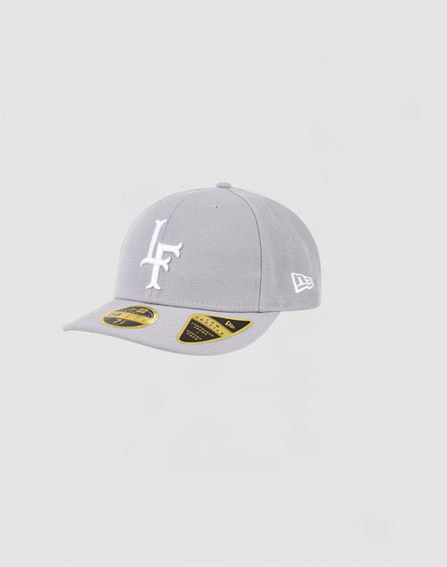 The LFDY x NEW ERA 59FIFTY Low Profile by LIVE FAST DIE YOUNG is a light grey fitted cap with a white L and F logo on the front, a white New Era logo on the side, and black and gold stickers on the brim.