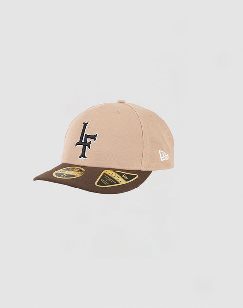 The LFDY x NEW ERA 59FIFTY Low Profile cap by LIVE FAST DIE YOUNG features a beige crown, brown brim, dark blue LF front logo, and white side logo. Fitted style with brim stickers, shown on a plain light gray background.