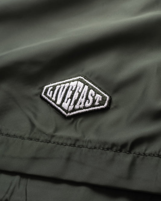 A close-up of olive green Basic Satin Shorts by LIVE FAST DIE YOUNG in polyester fabric features a white and black embroidered logo patch and a comfortable elastic waistband.
