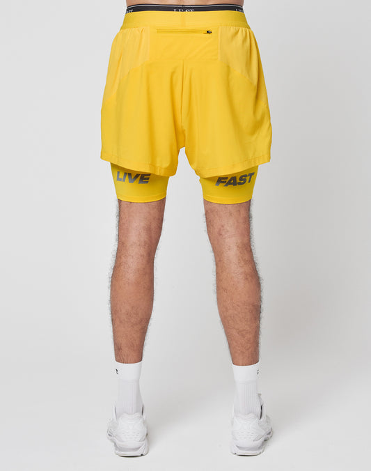 A person stands facing away, wearing LIVE FAST DIE YOUNG’s Live Fast Performance Running Shorts with integrated tights and branding on the legs, paired with white socks and sneakers against a plain background.