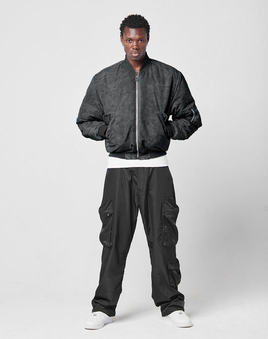 A person stands against a plain light gray background wearing the LIVE FAST DIE YOUNG Washed Bomber Jacket, black cargo pants, a white shirt, and white sneakers. Their hands are in their jacket pockets.