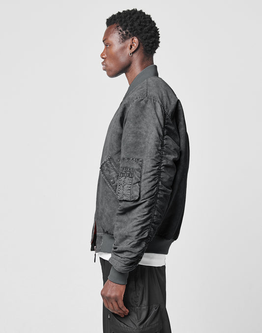 A man stands in profile against a plain background, wearing the LIVE FAST DIE YOUNG Washed Bomber Jacket with visible stitching and sleeve pockets, layered over a white shirt and black pants.