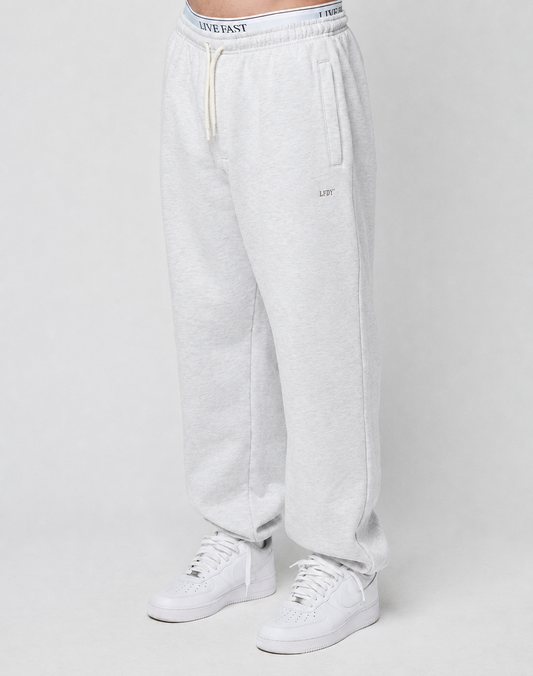 A person wears the LIVE FAST DIE YOUNG Basic Loose Jogger in light gray, featuring a logo print waistband with white drawstring, side pockets, and small embroidered detail on the left thigh, paired with white sneakers against a plain background.