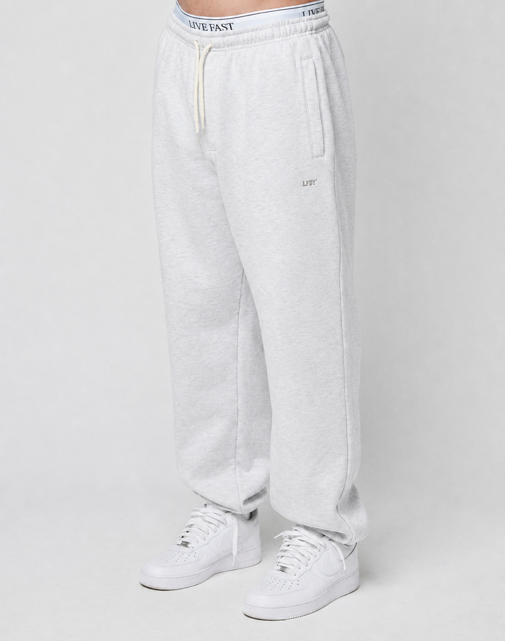 A person wears the LIVE FAST DIE YOUNG Basic Loose Jogger in light gray, featuring a logo print waistband with white drawstring, side pockets, and small embroidered detail on the left thigh, paired with white sneakers against a plain background.