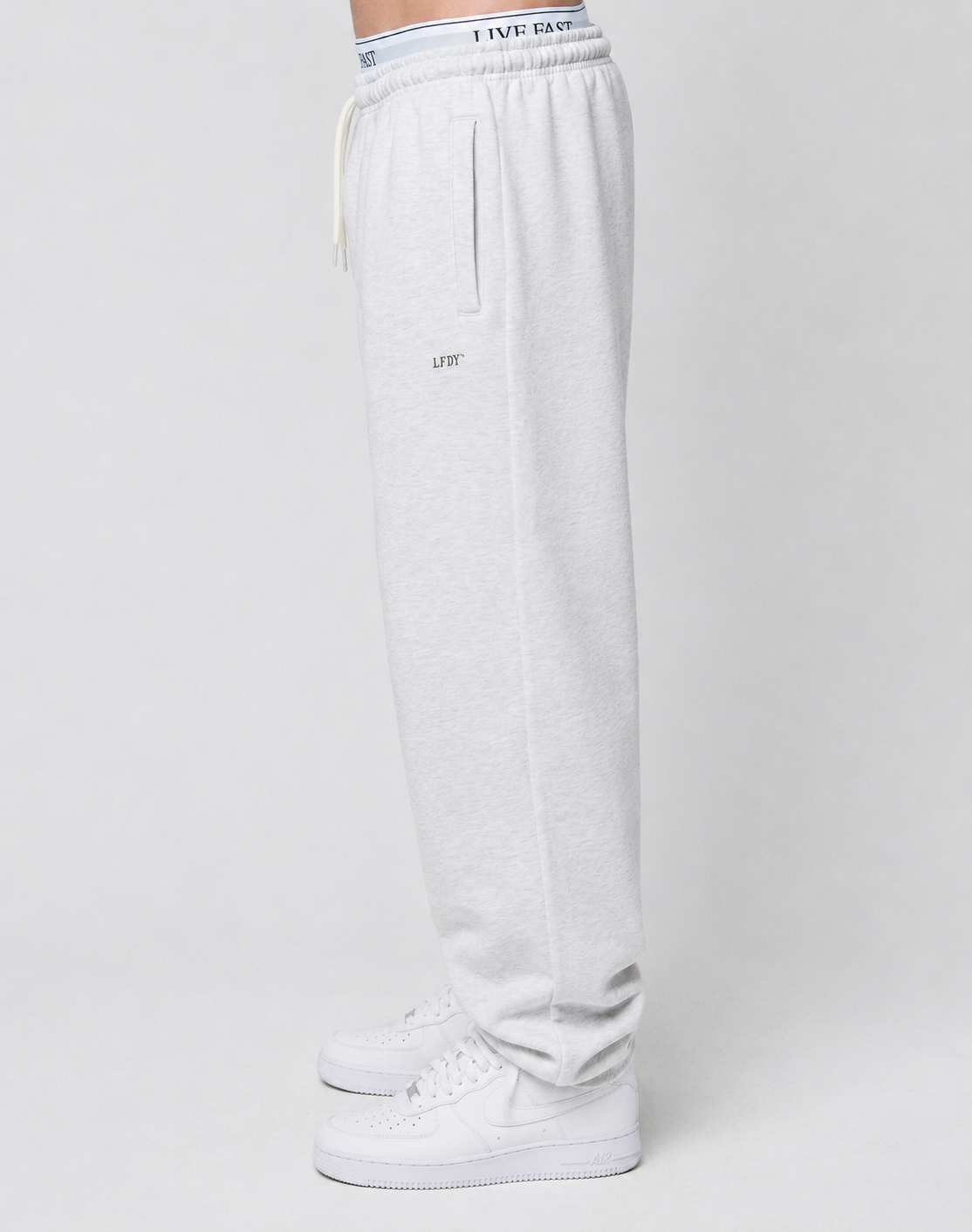 A person stands sideways in Basic Loose Jogger sweatpants from LIVE FAST DIE YOUNG, featuring a relaxed fit and white sneakers. The Elastik-Bund with the LIVE FAST logo is visible above the pants against a plain light background.
