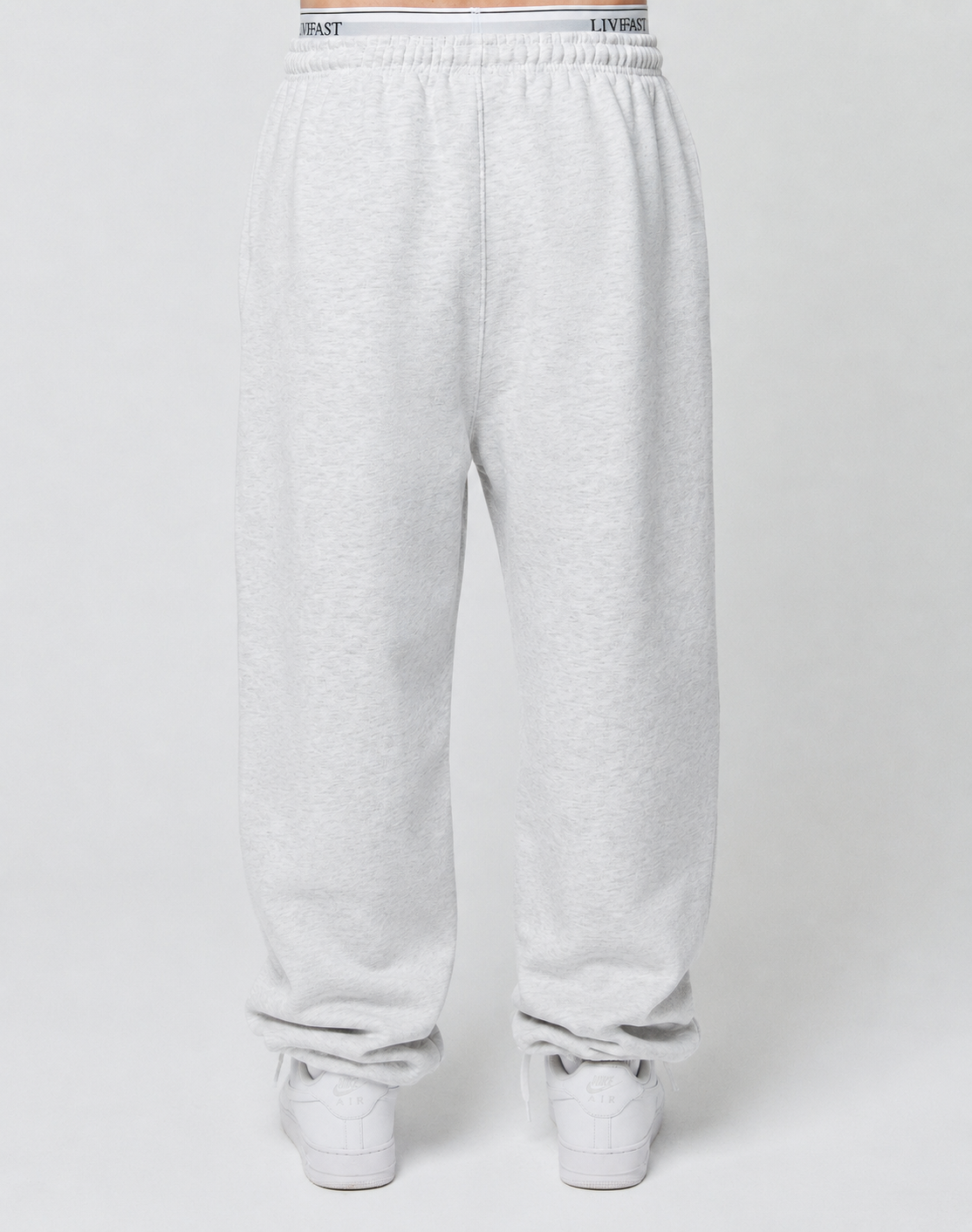A person wears LIVE FAST DIE YOUNG Basic Loose Jogger sweatpants in light gray with an elastic waistband and loose fit, paired with white sneakers, standing against a plain white background. The joggers look comfortable and relaxed.