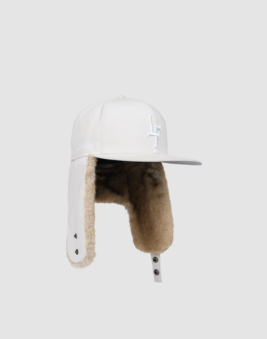 The LFDY x New Era 59Fifty DogEar by LIVE FAST DIE YOUNG is a white fitted cap with tan faux fur ear flaps, interior lining, a white embroidered front logo, and a stud chin closure, shown on a light gray background.
