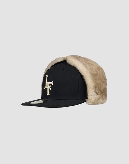 The LFDY x New Era 59Fifty DogEar by LIVE FAST DIE YOUNG features beige faux fur ear flaps, a gold LF logo on the front, and the classic New Era Flag, shown against a plain light gray background.
