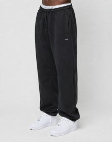 A person is wearing the Basic Loose Jogger by LIVE FAST DIE YOUNG, featuring black fabric, an Elastik-Bund drawstring waist, a visible white LIVE FAST waistband, and white sneakers, standing against a plain light background.