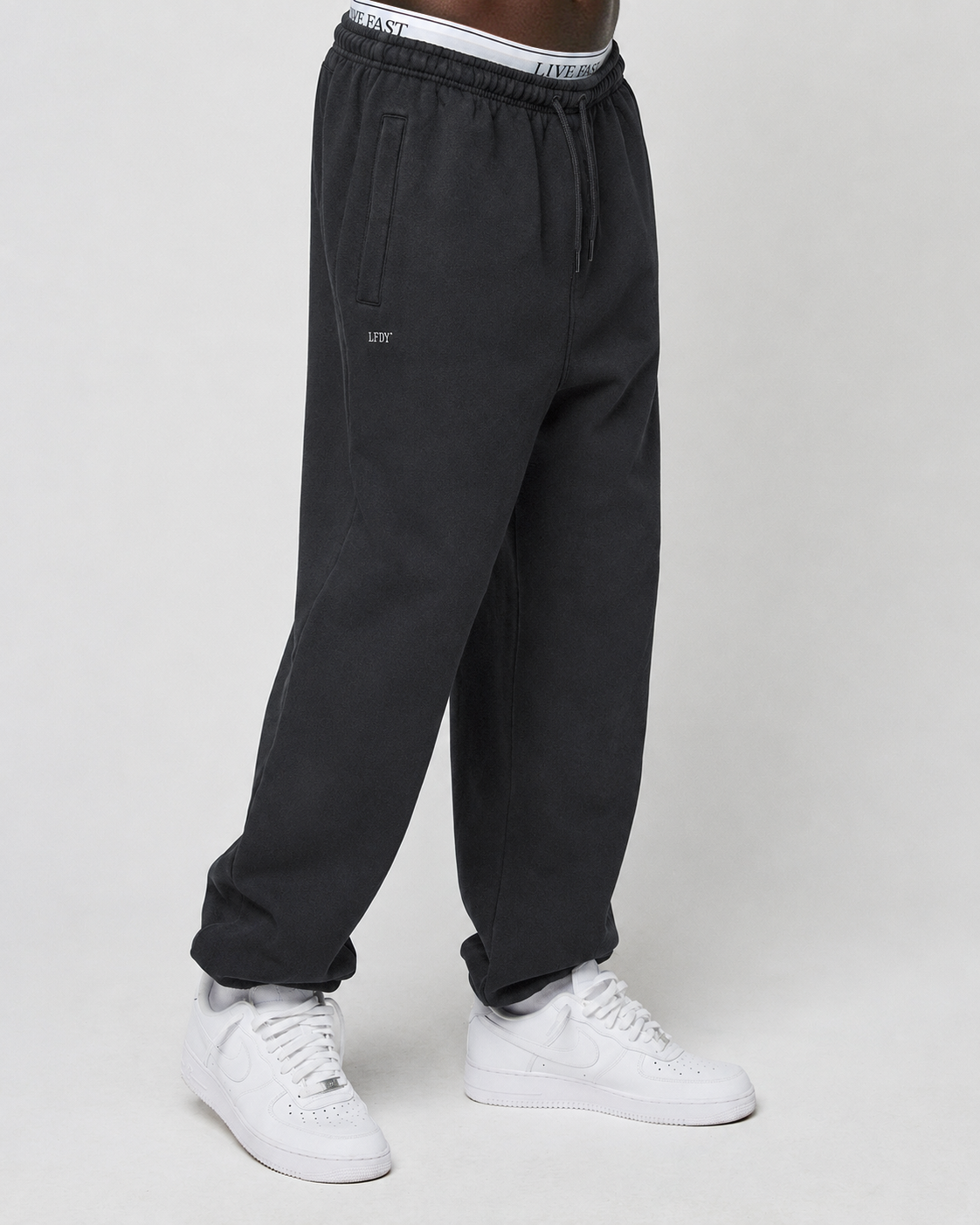 A person wears the LIVE FAST DIE YOUNG Basic Loose Jogger—black sweatpants with an elastik-bund and logo print waistband—paired with white sneakers, set against a plain light grey background.