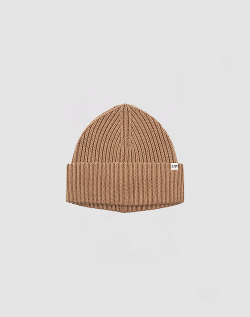 The LIVE FAST DIE YOUNG Rib Knit Beanie is tan, ribbed, one size, with a folded cuff and a small white flag label on the side, shown against a plain white background.