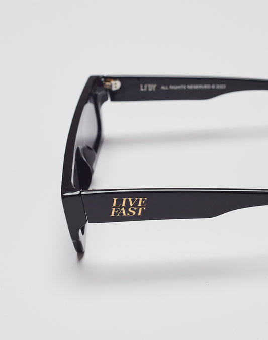 The Live Fast Shades by LIVE FAST DIE YOUNG are black sunglasses with LIVE FAST in gold on the temple, made from polished LAES acetate, offer 400 UV protection, and are shown against a plain white background.