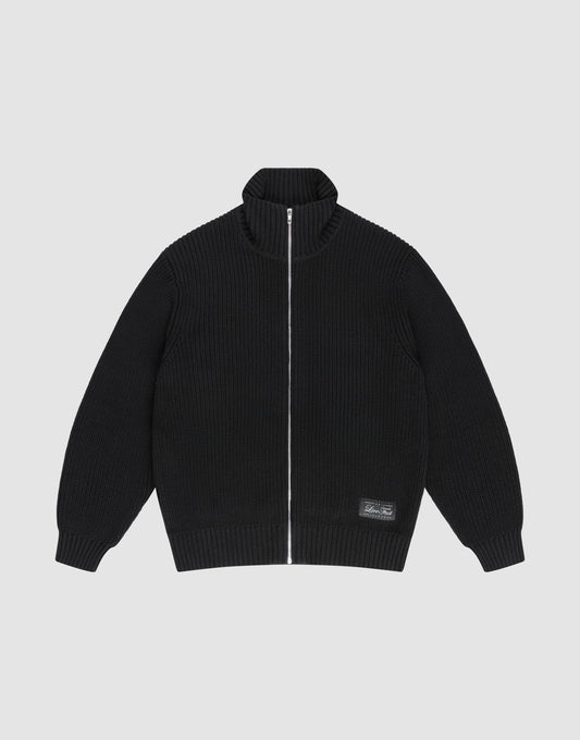 The LFDY Knit Jacket by LIVE FAST DIE YOUNG is a black, ribbed zip-up with Stehkragen, long sleeves, and a small rectangular patch near the hem. Made from 100% Baumwolle, it's shown laid flat on a light gray background. | plain
