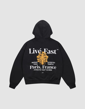 The LFDY Paris Hooded Zip by LIVE FAST DIE YOUNG is an oversized black cotton hoodie with front and back prints: features white “Live Fast” and “Paris, France” text, a gold floral cross graphic, small white text, and a street address. | plain