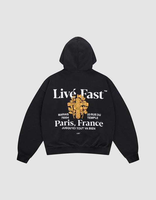 The LFDY Paris Hooded Zip by LIVE FAST DIE YOUNG is an oversized black cotton hoodie with front and back prints: features white “Live Fast” and “Paris, France” text, a gold floral cross graphic, small white text, and a street address. | plain