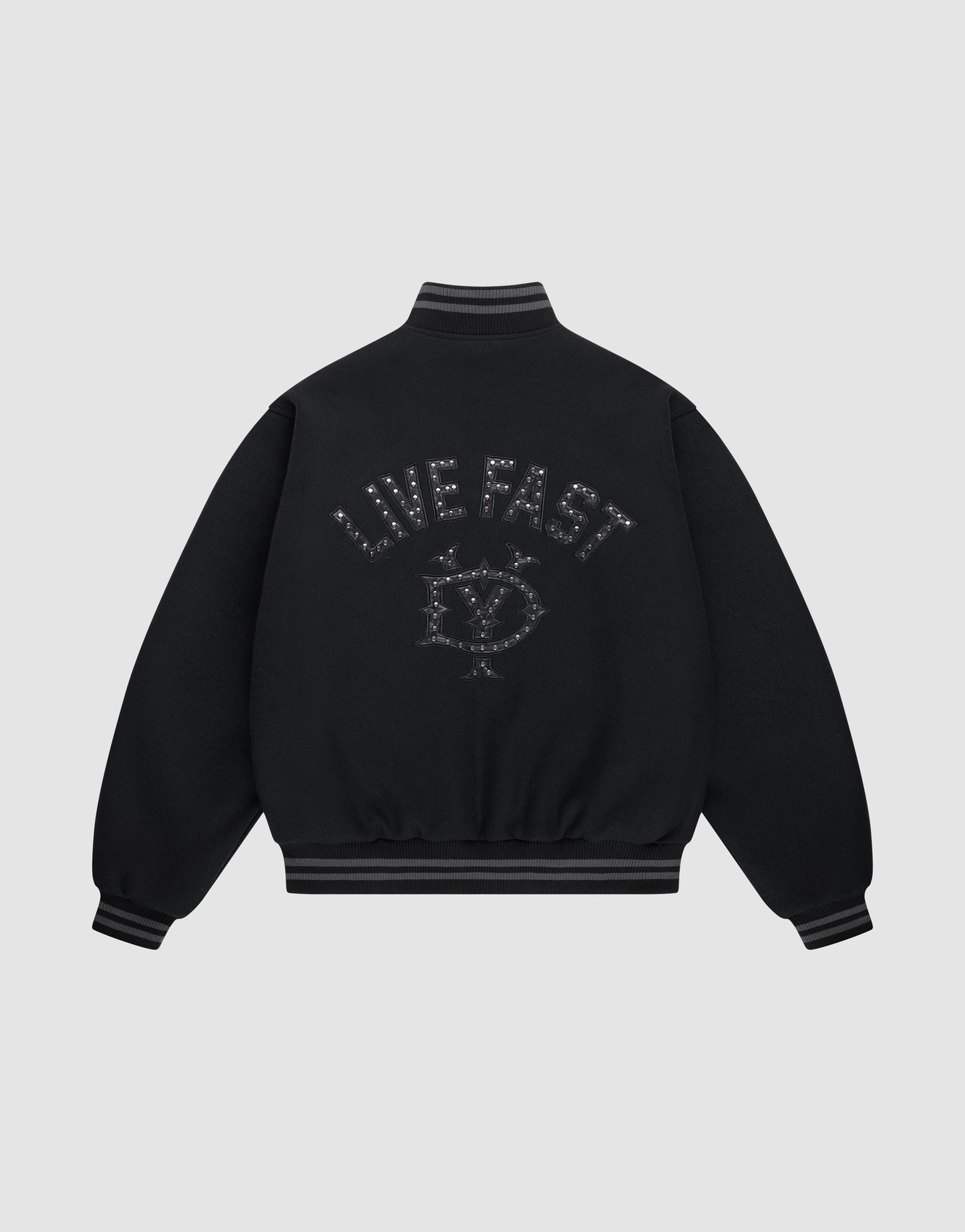 The Live Fast College Jacket by LIVE FAST DIE YOUNG is a black varsity jacket with striped cuffs and hem, a metal zipper, and an embroidered eye with stars and "LIVE FAST" on the back in dark thread. The background is light gray.