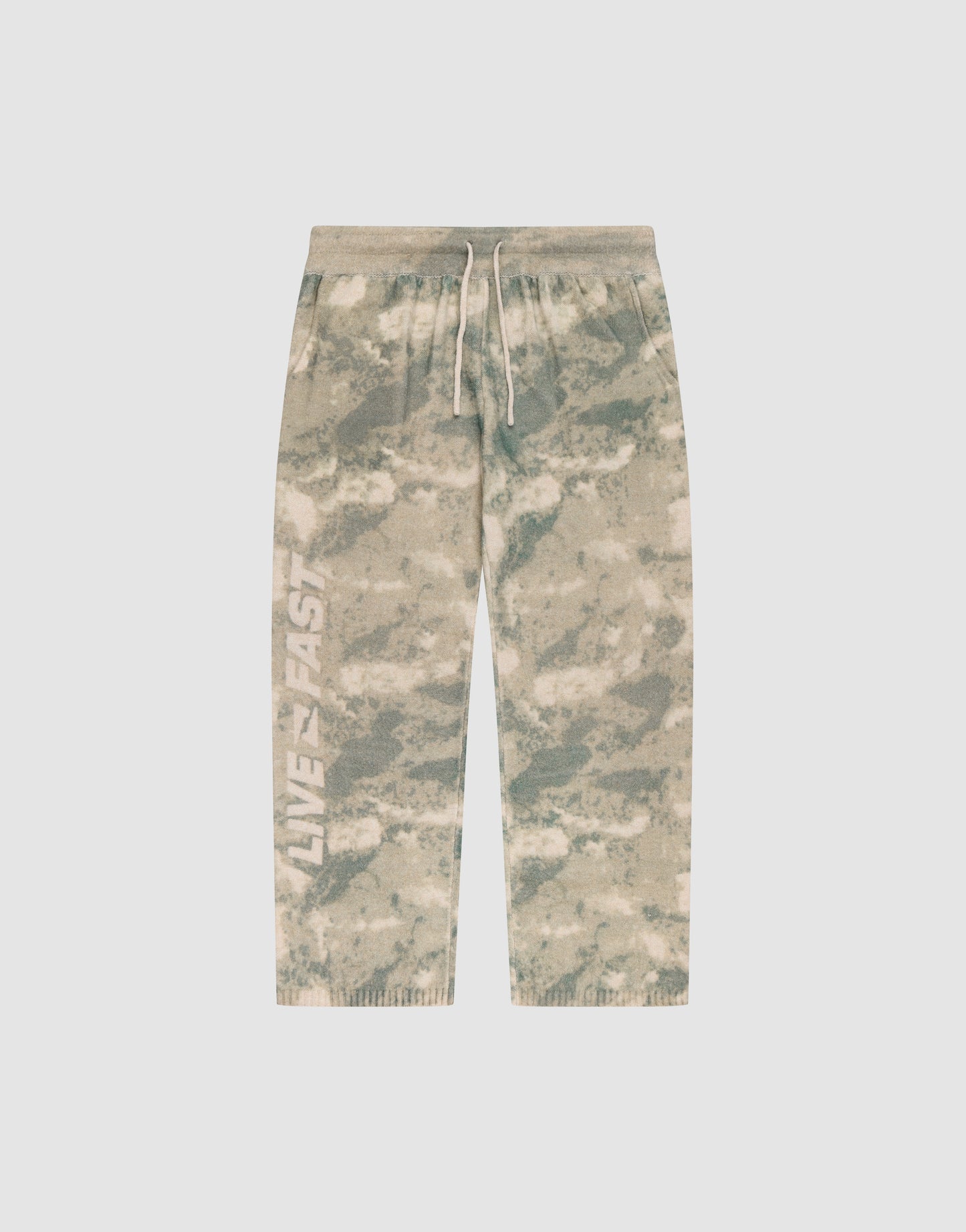 The LIVE FAST DIE YOUNG Live Fast Boucle Pants feature a beige and green tie-dye design, white drawstring, elastic waistband, "LIVE FAST" on the left leg, relaxed wide fit with straight leg and ribbed cuffs for comfort.