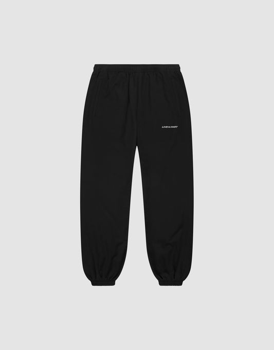 LFDY Trackpants by LIVE FAST DIE YOUNG: black regular-fit joggers with elastic waistband and cuffs, side pockets, and a small white logo on the upper left thigh, shown on a plain light grey background. | plain