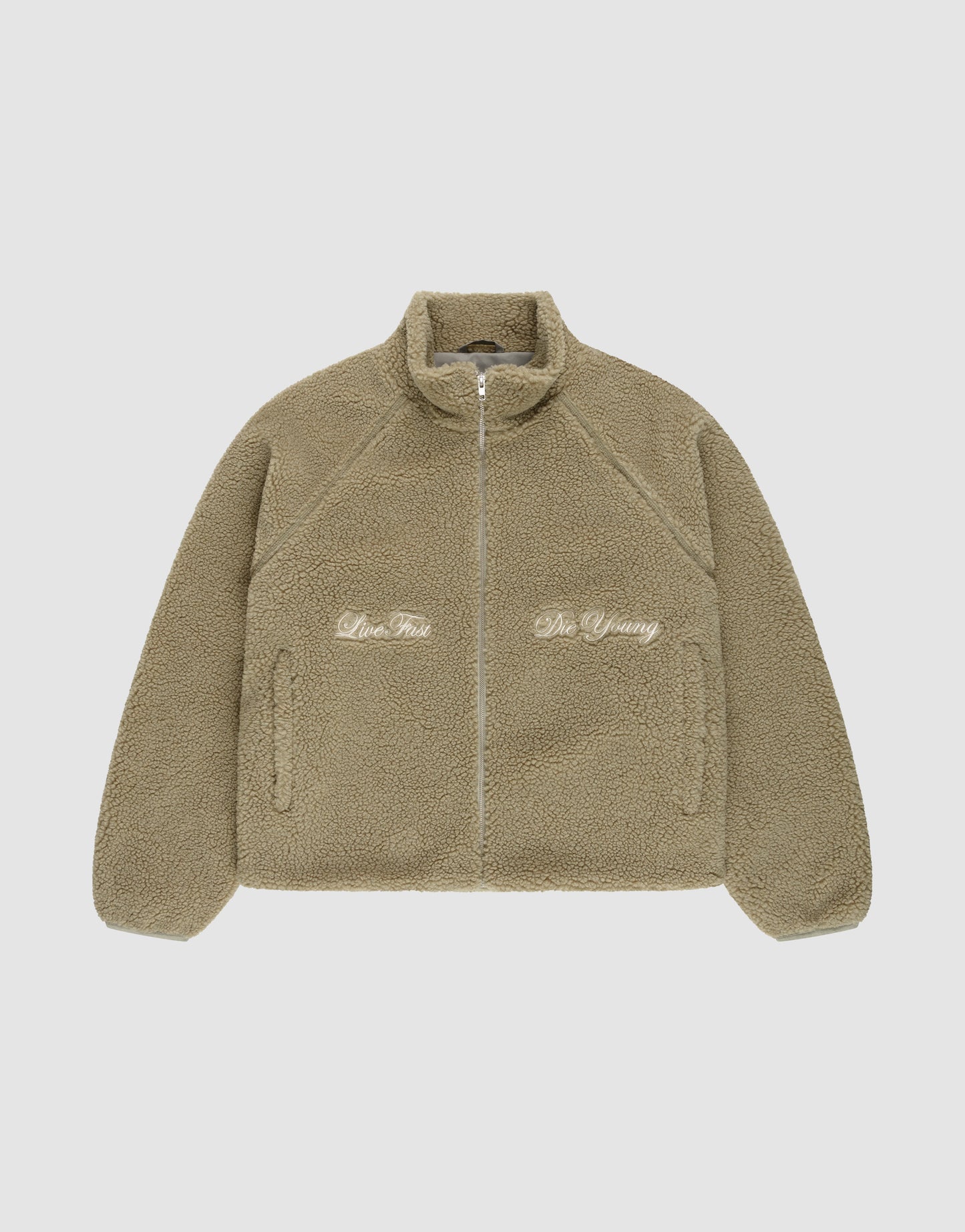 The LFDY Teddy Jacket by LIVE FAST DIE YOUNG is a tan oversized fleece jacket with a high collar and raglan sleeves. Embroidered text "Live Fast" appears on the left and "Die Young" on the right, shown laid flat on a white background.
