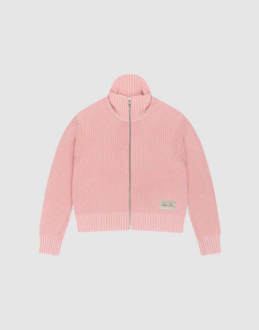 The LFDY Knit Jacket by LIVE FAST DIE YOUNG is a light pink, ribbed zip-up sweater with a stand collar and long sleeves, made from 100% cotton. A small beige patch accents the lower left front. Shown here laid flat on a white background. | plain