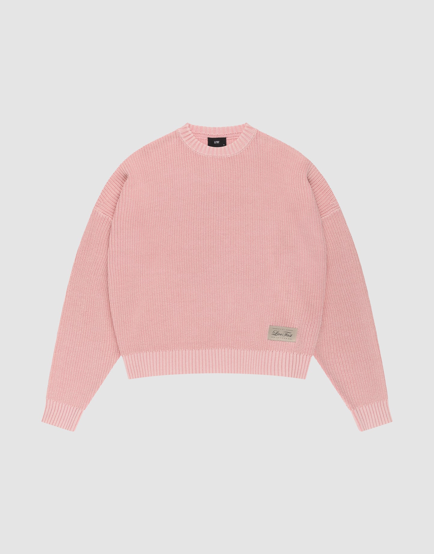 The LFDY Knit Sweater by LIVE FAST DIE YOUNG features pink ribbed knit, an oversized fit, dropped shoulders, long sleeves, a crew neckline, and a small rectangular beige patch with text on the front hem, displayed on a plain white background.