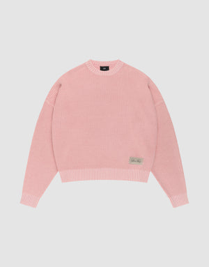 The LFDY Knit Sweater by LIVE FAST DIE YOUNG features pink ribbed knit, an oversized fit, dropped shoulders, long sleeves, a crew neckline, and a small rectangular beige patch with text on the front hem, displayed on a plain white background. | plain