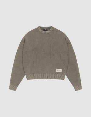 The LFDY Knit Sweater by LIVE FAST DIE YOUNG is a ribbed, oversized crewneck in muted olive green Baumwolle with long sleeves, dropped shoulders, and a small cream text patch on the front left. Shown flat on a light gray background. | plain