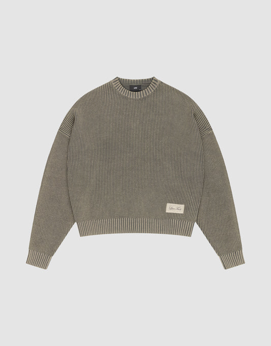The LFDY Knit Sweater by LIVE FAST DIE YOUNG is a ribbed, oversized crewneck in muted olive green Baumwolle with long sleeves, dropped shoulders, and a small cream text patch on the front left. Shown flat on a light gray background. | plain