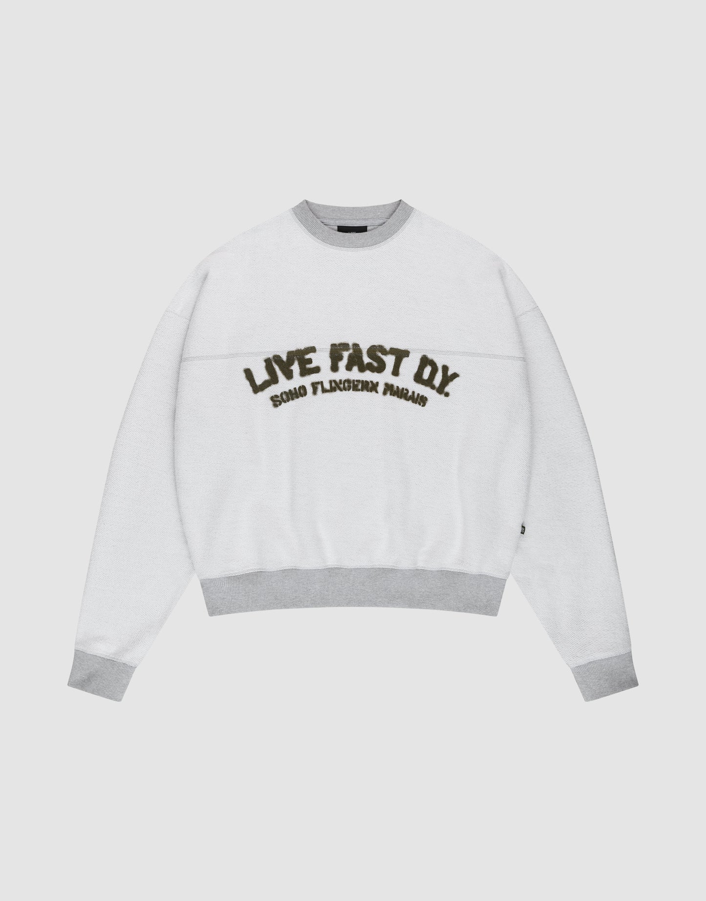 The LFDY District Sweater by LIVE FAST DIE YOUNG is a white oversized crewneck with light gray collar, cuffs, and hem in 480 GSM inside-out fabric. The chest displays bold LIVE FAST D.I.Y. text above ROAD FLECHTERIN PARADIS.
