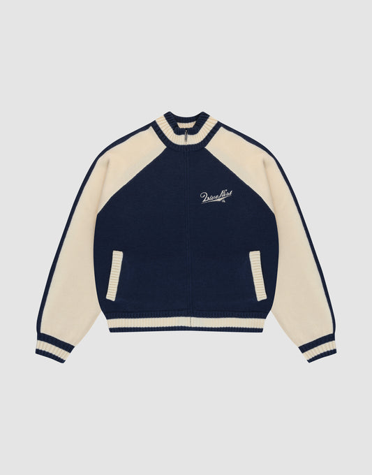 The LIVE FAST DIE YOUNG Boxy Knit Zip Jacket features a navy and cream varsity style with cream raglan sleeves, striped cuffs and collar, two front pockets, and Paradise embroidery on the left chest. Set against a plain light gray background.