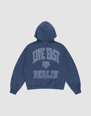 The LIVE FAST DIE YOUNG Live Fast Berlin Hooded Zip is an oversize fit blue sweatshirt with a hood, featuring "LIVE FAST" in large letters, a small graphic below, and bold "BERLIN" on the back. Made from 100% Baumwolle with a vintage wash. Shown on white background. | plain