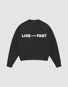 The Live – Fast Sweater by LIVE FAST DIE YOUNG is a black, oversized crewneck cotton pullover with "LIVE — FAST" in bold white glossy silicone print on the chest, shown laid flat against a plain light gray background. | plain