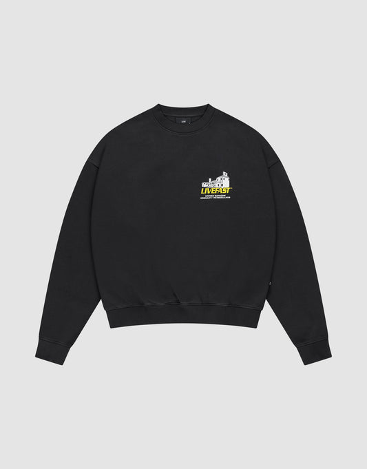 The LFDY Supply Sweater by LIVE FAST DIE YOUNG is a black oversized crewneck with a small colorful graphic and “LIVEST at the university” text on the left chest, crafted from a soft cotton-polyester blend and shown on a light grey background. | plain