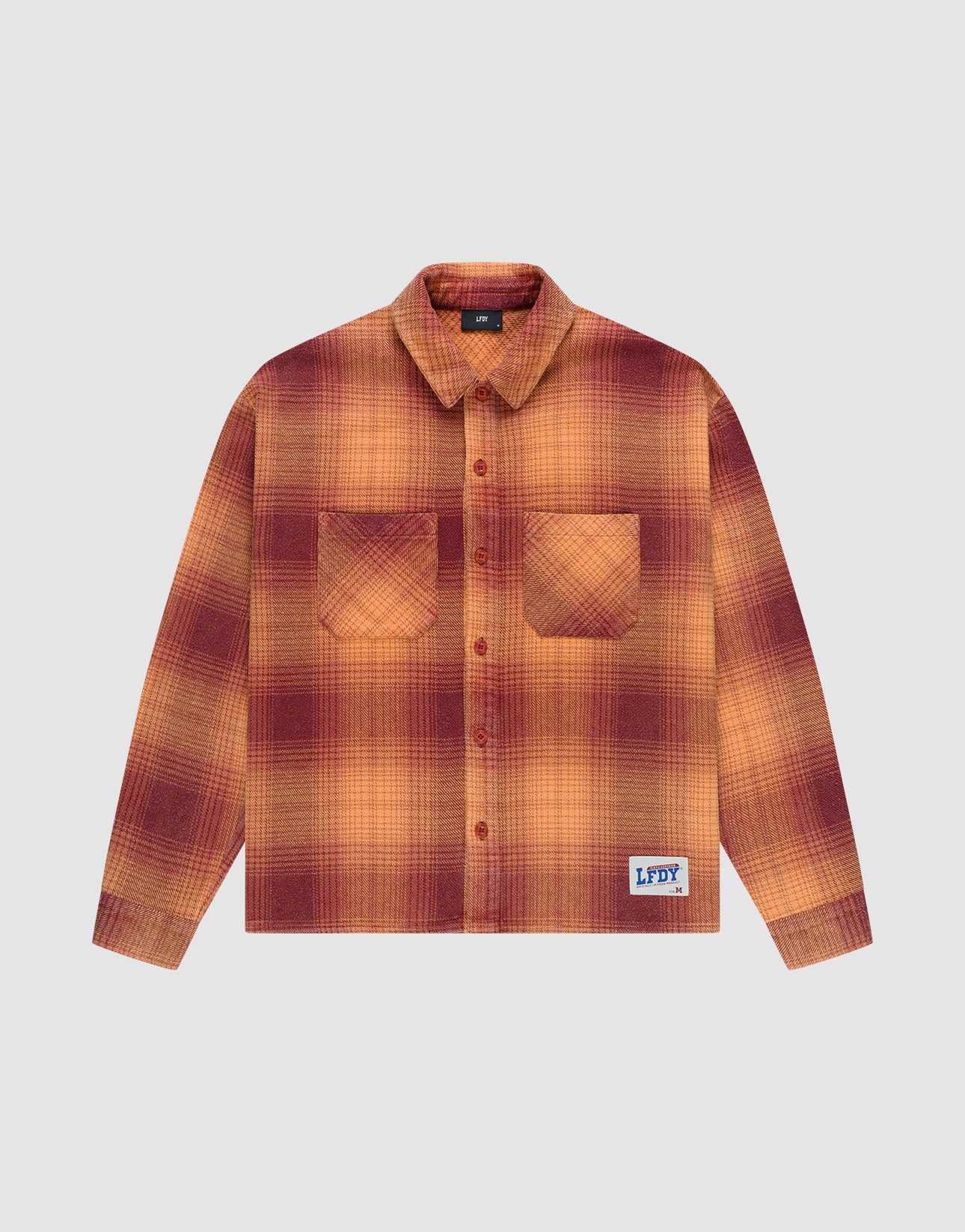 The LIVE FAST DIE YOUNG Vintage Flannel is a long-sleeve, button-up shirt with an oversized fit, red and orange plaid pattern, two chest pockets, and a blue brand patch near the hem, shown on a plain light background.
