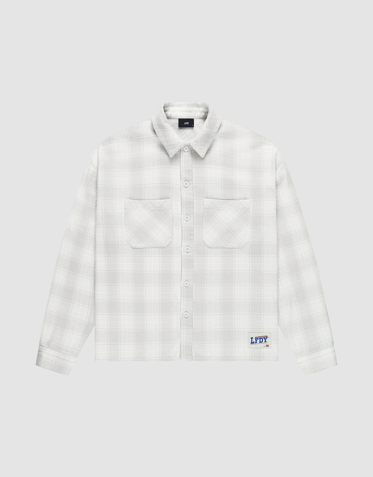 The LIVE FAST DIE YOUNG Vintage Flannel is a white plaid oversize long-sleeve shirt with two chest pockets, vintage wash, and a blue logo patch above the hem on a light grey background.