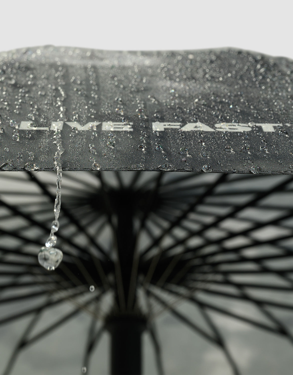 Close-up of the black LFDY Umbrella by LIVE FAST DIE YOUNG, water droplets on its surface and one falling off the edge. The underside and metal struts highlight its stable frame, with the brand text partly visible through the drops.