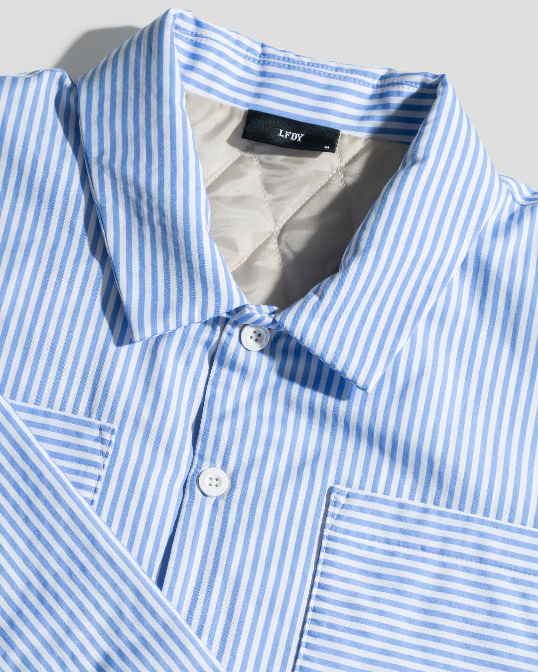 A close-up of the LFDY Striped Padded Shirt by LIVE FAST DIE YOUNG, featuring blue and white vertical stripes, a collar, Brusttaschen, and a front pocket, displayed on a flat white background.