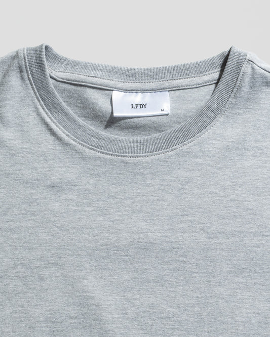 A close-up of the LFDY Slim Tee by LIVE FAST DIE YOUNG in light grey, featuring a white neck label with brand and size M, plus a signature Flag-Label. The slim fit T-shirt is shown laid flat on a light background.