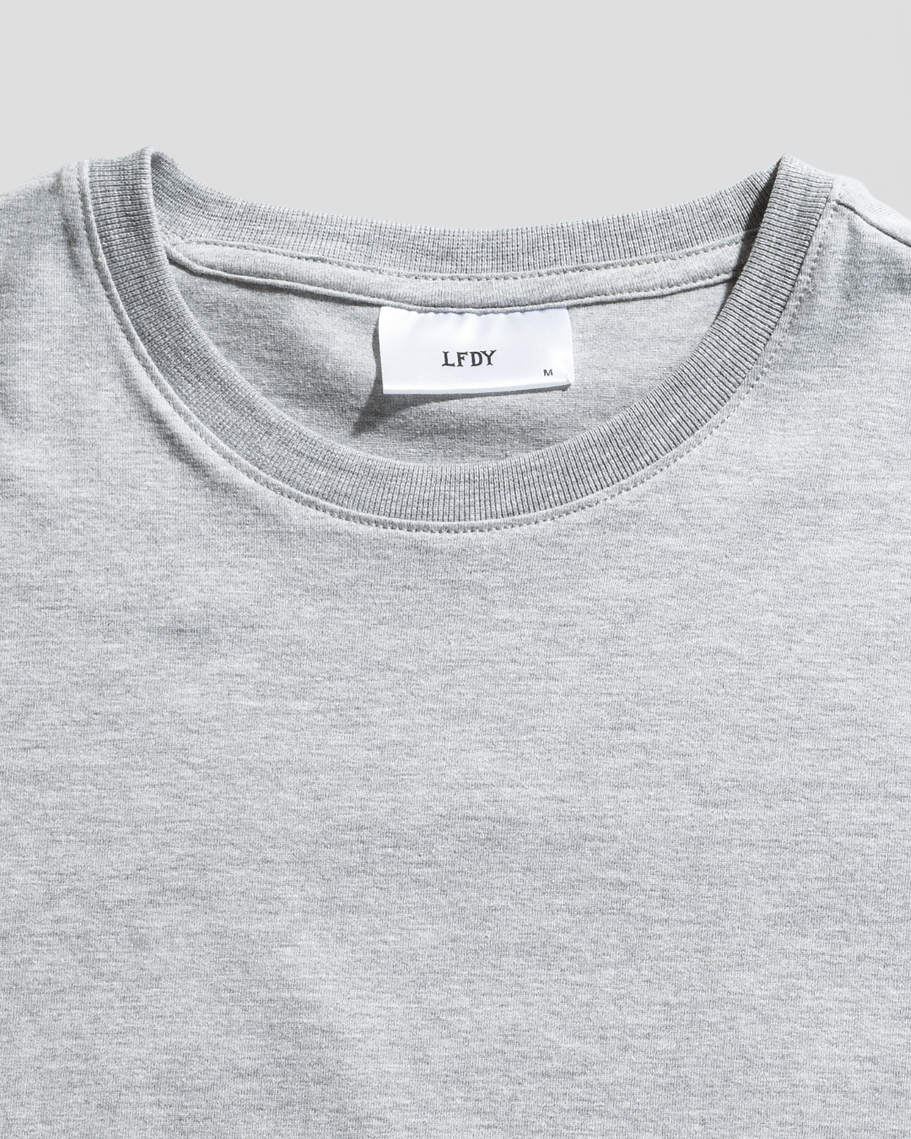 A close-up of the LFDY Slim Tee by LIVE FAST DIE YOUNG in plain light gray, featuring a collar with a white neck tag showing LFDY, size M, Flag-Label details, and crafted from soft Baumwolle.
