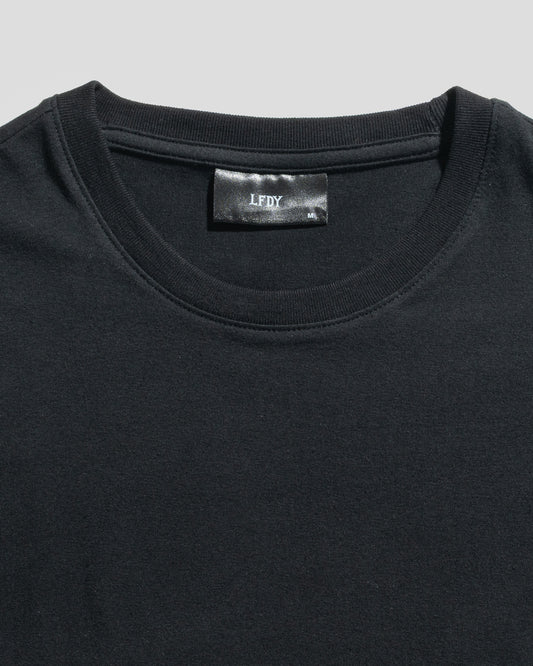 Close-up of a plain black LFDY Slim Tee by LIVE FAST DIE YOUNG, lying flat to reveal the upper chest and neckline. A black label with "LFDY" text is visible inside the collar. The shirt is crafted from soft cotton.