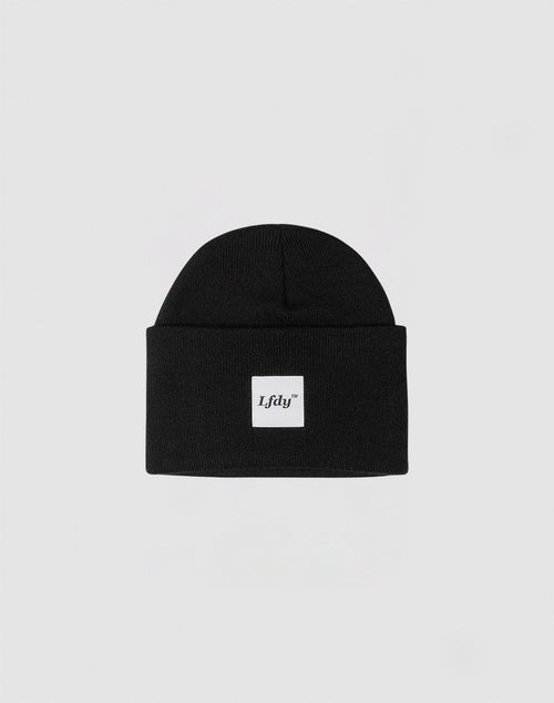 The LFDY Labeled Beanie is a black polyacryl knit with a folded brim and a small white rectangular Idfy logo patch on the front. Shown on a plain light gray background.