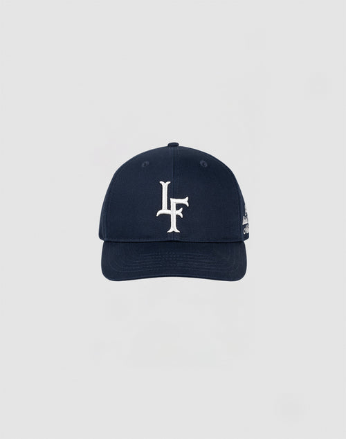 The LF Snapback by LIVE FAST DIE YOUNG is a navy blue cotton baseball cap with a large white LF embroidered in front and a small logo on the side, displayed against a plain light gray background.