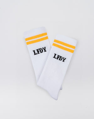 The LFDY Logo Socks (2 Pack) by LIVE FAST DIE YOUNG feature a white ribbed design with two yellow stripes and black LFDY text below. Made from 82% cotton, they’re easy-care—please follow the care instructions.
