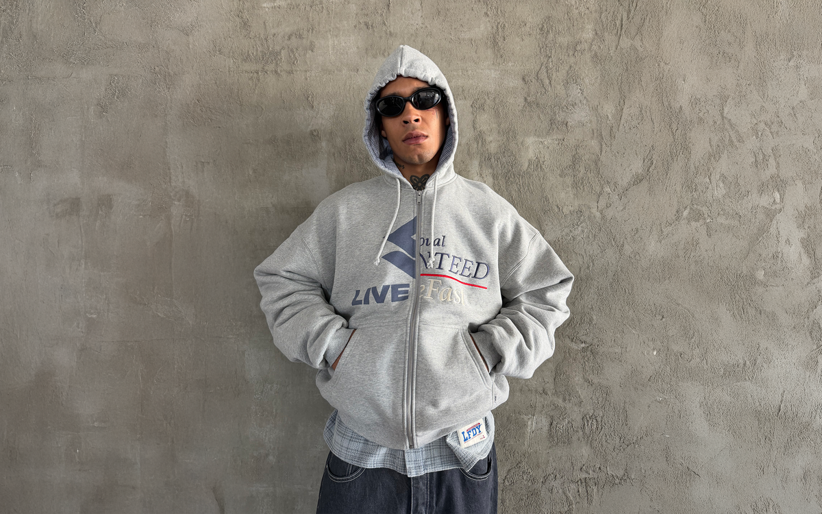 Person wearing a gray hooded zip-up sweatshirt, sunglasses, and layered clothing stands with hands in pockets against a textured gray concrete wall.